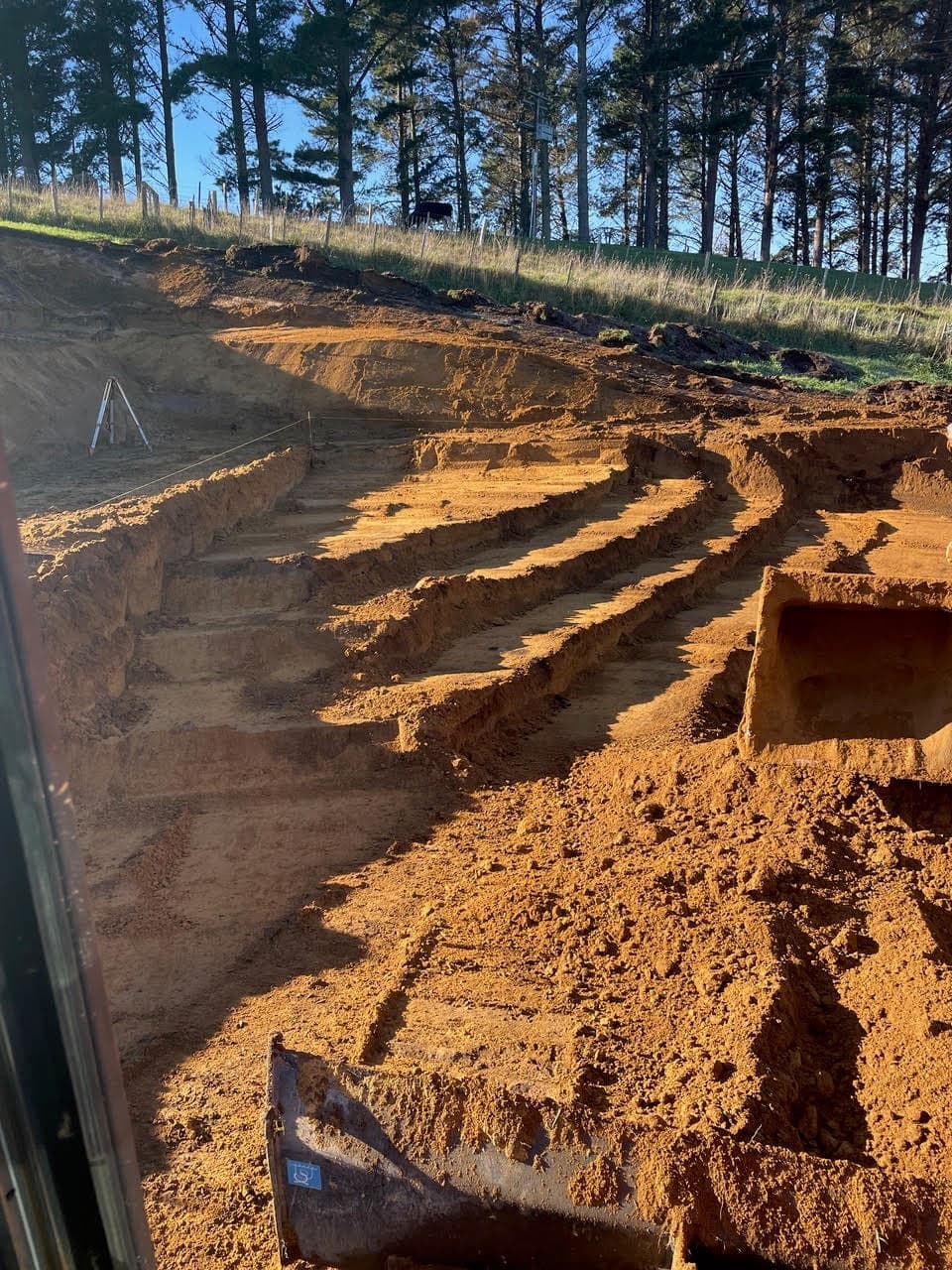 Stepped benches cut into a clay bank, seen from the digger cab with the laser level set up above
