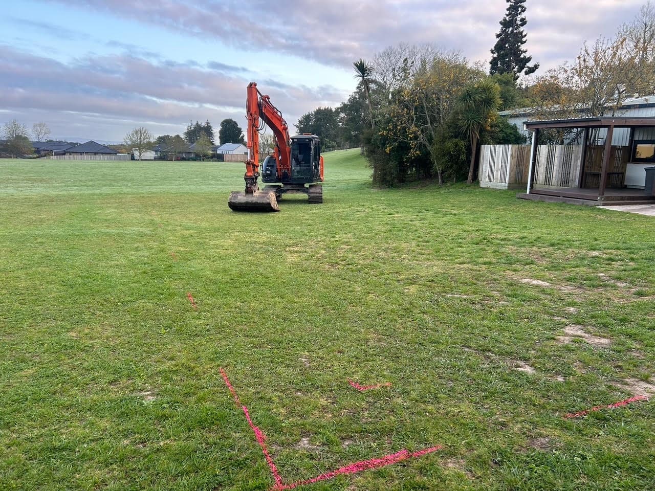 Orange digger tracking onto a grassed section beside a house, the layout marked out in paint