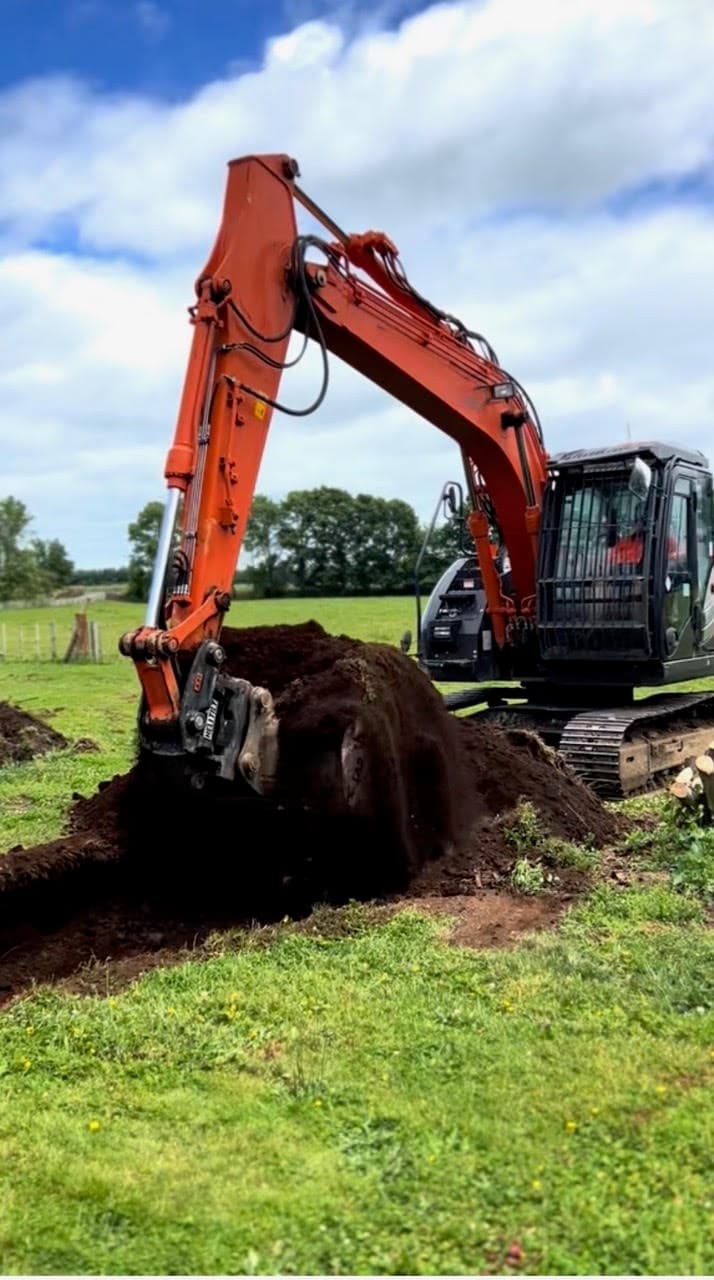 Orange digger with its bucket in the ground, digging in a green paddock