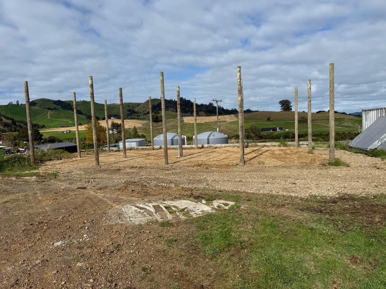 Pole shed poles standing on a metalled pad, grain silos and rolling Waikato hills behind