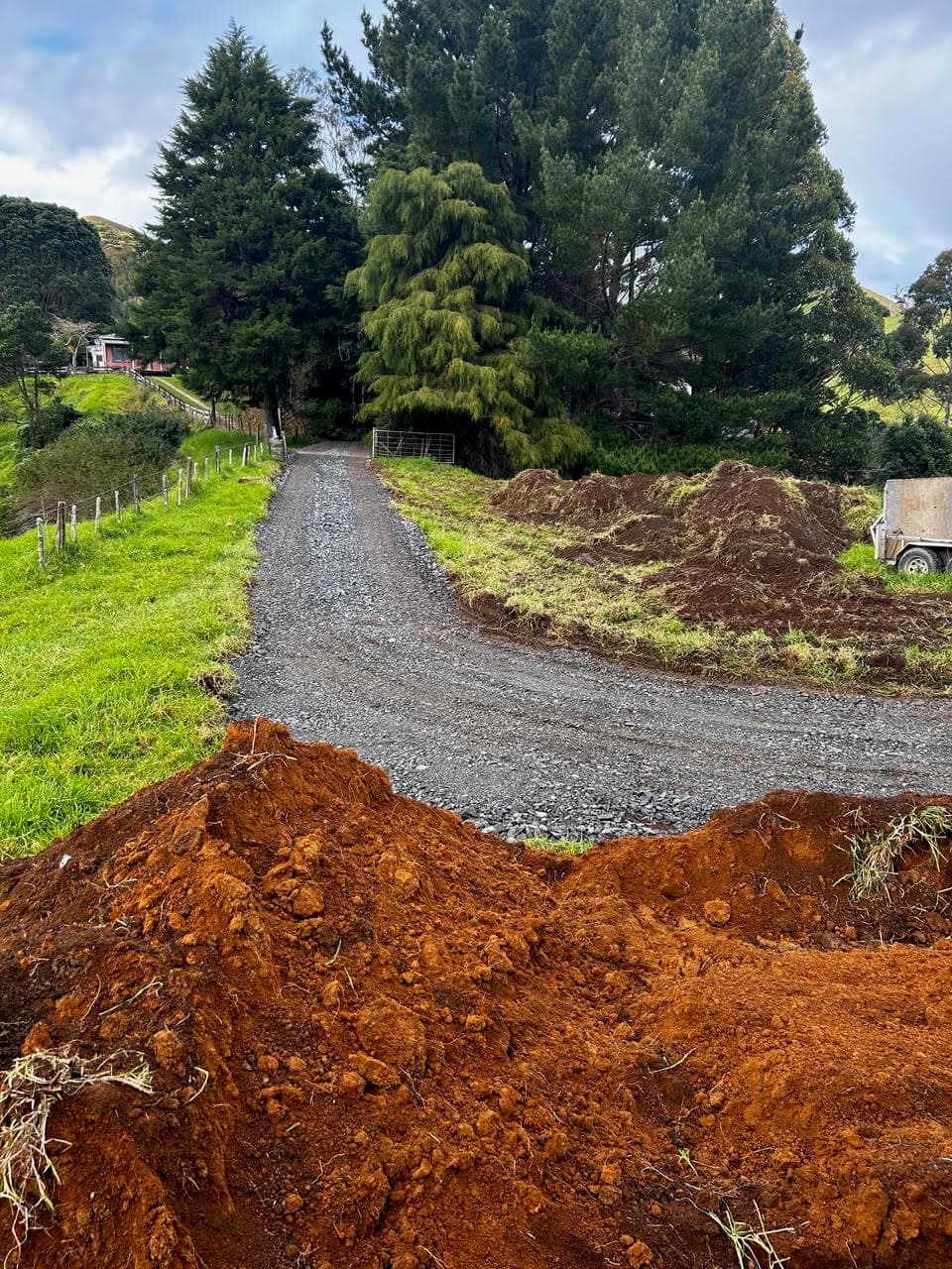 Freshly metalled rural driveway running up to the gate under tall trees, dug clay heaped in the foreground